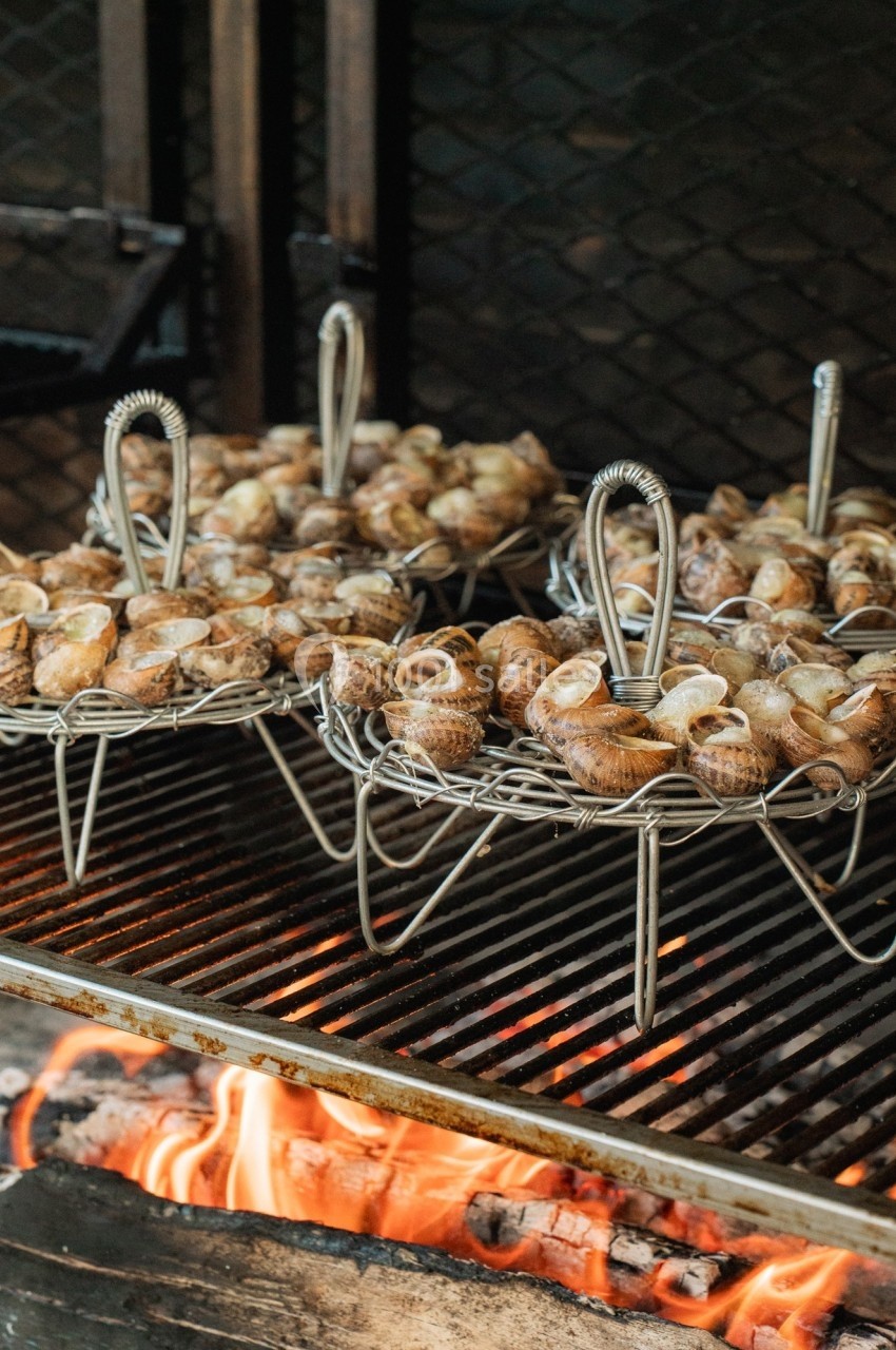 Escargots disposés dans des paniers métalliques, grillant sur un barbecue au-dessus de flammes vives.