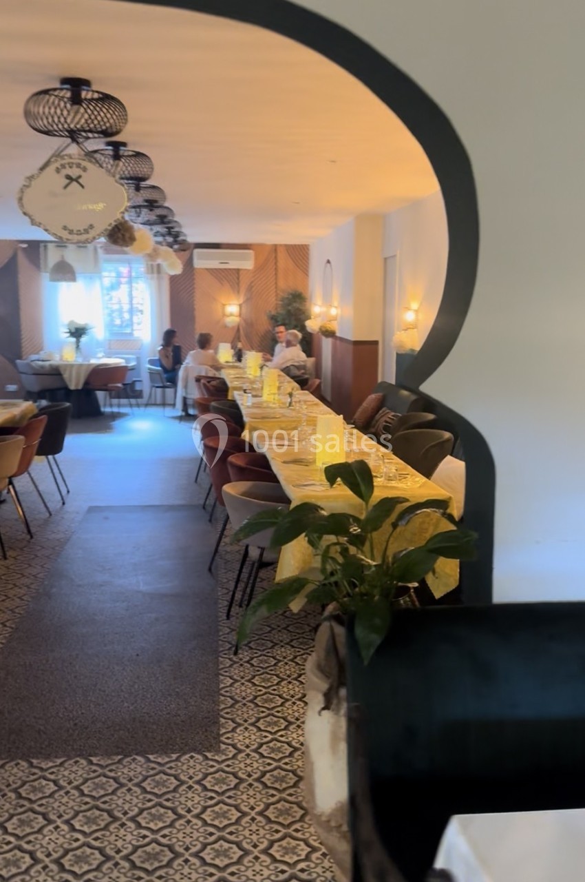 Salle de restaurant lumineuse avec une longue table dressée, décorée de plantes et éclairée par des suspensions.