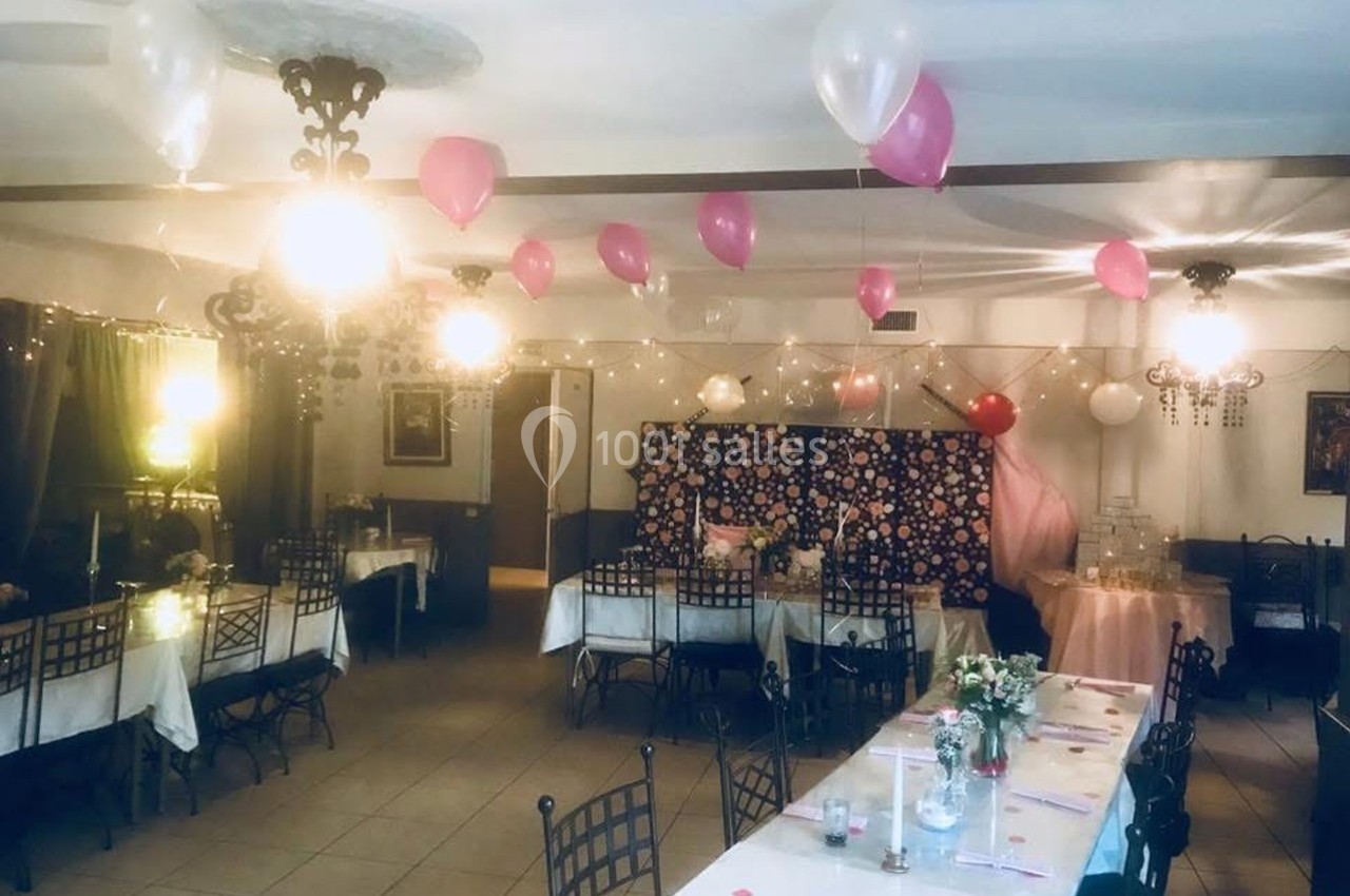 Salle décorée pour une réception avec ballons roses et blancs, guirlandes lumineuses et tables dressées.