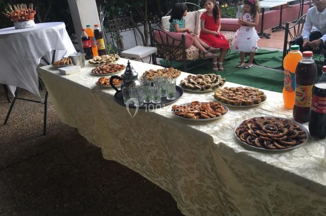 Table garnie de pâtisseries variées, théière avec verres, et boissons, en extérieur avec des enfants assis en arrière-plan.
