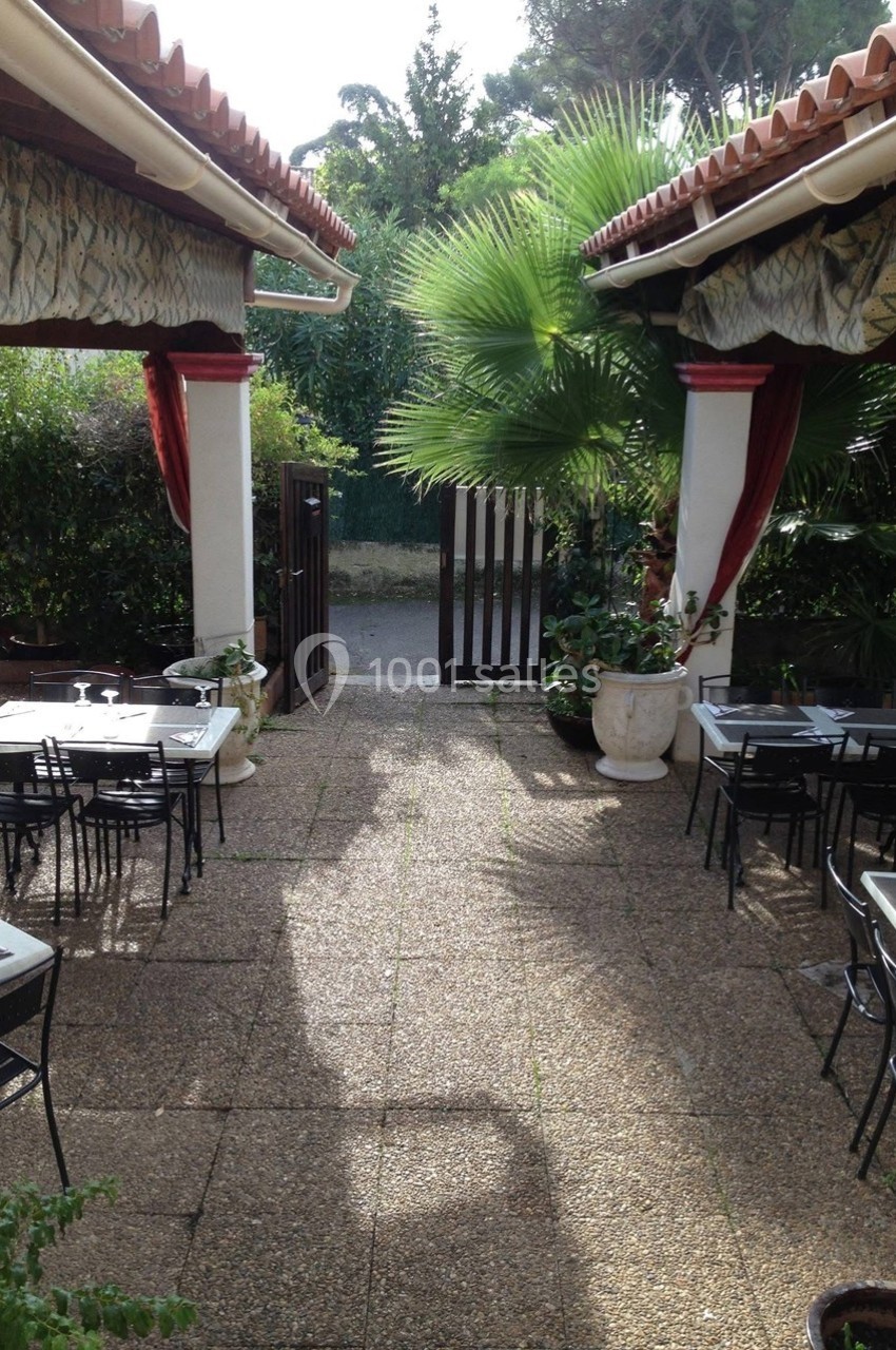 Terrasse extérieure avec tables et chaises en métal, entourée de plantes et ouverte sur un portail en bois.