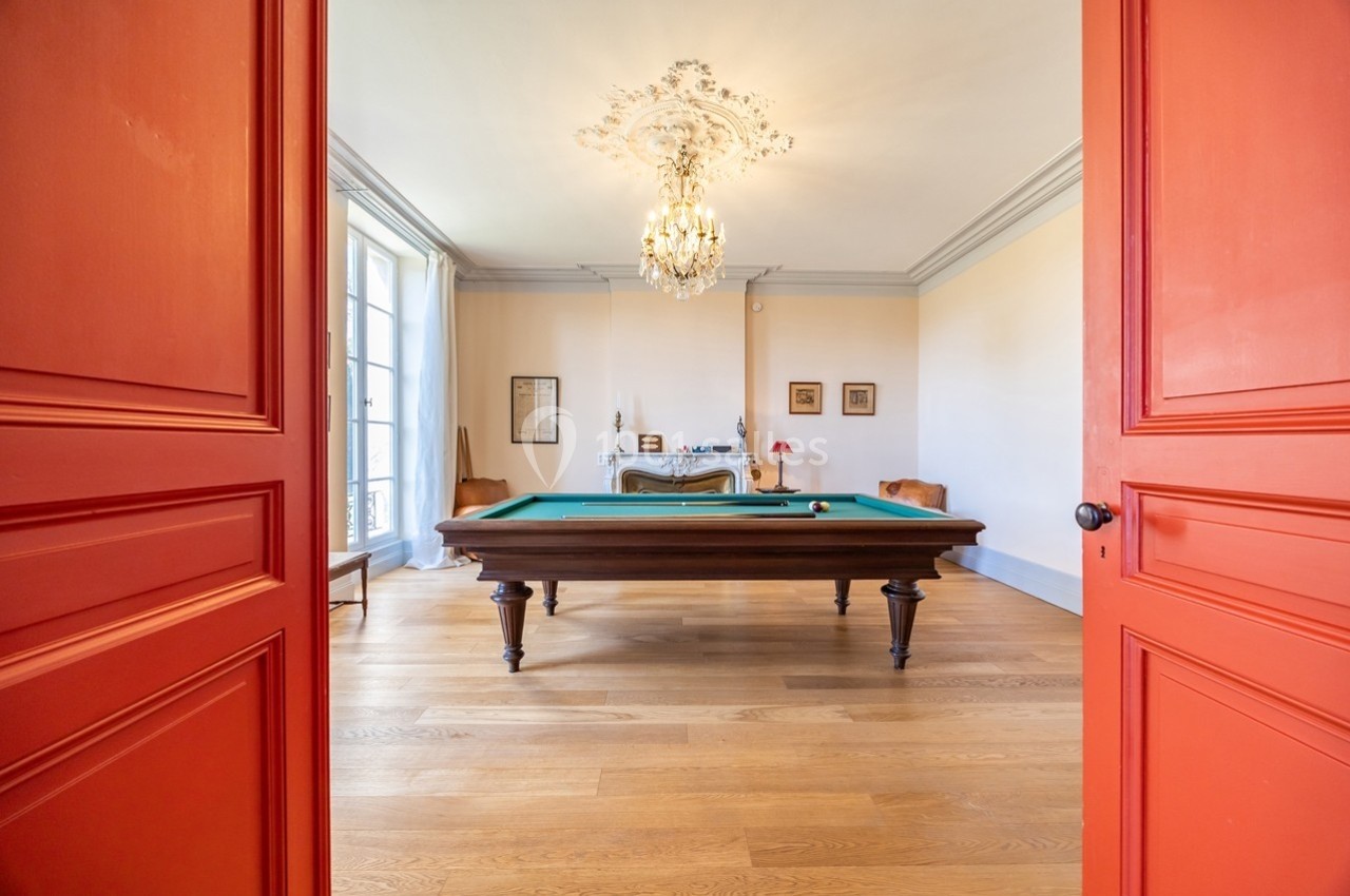 Salle avec table de billard, parquet en bois, murs clairs, lustre élégant et portes rouges ouvertes.