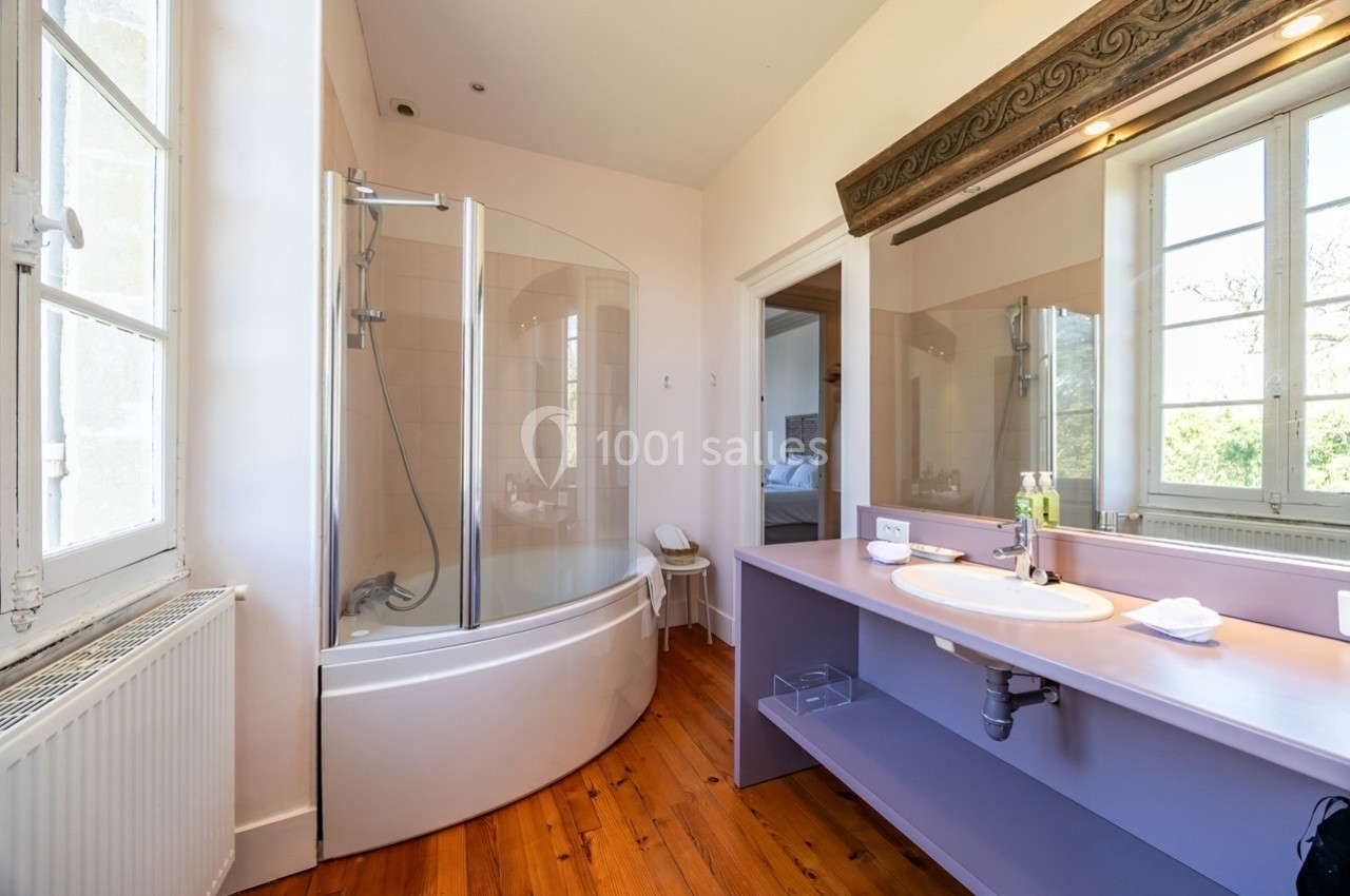 Salle de bain lumineuse avec baignoire d'angle, lavabo sur meuble violet et parquet en bois.