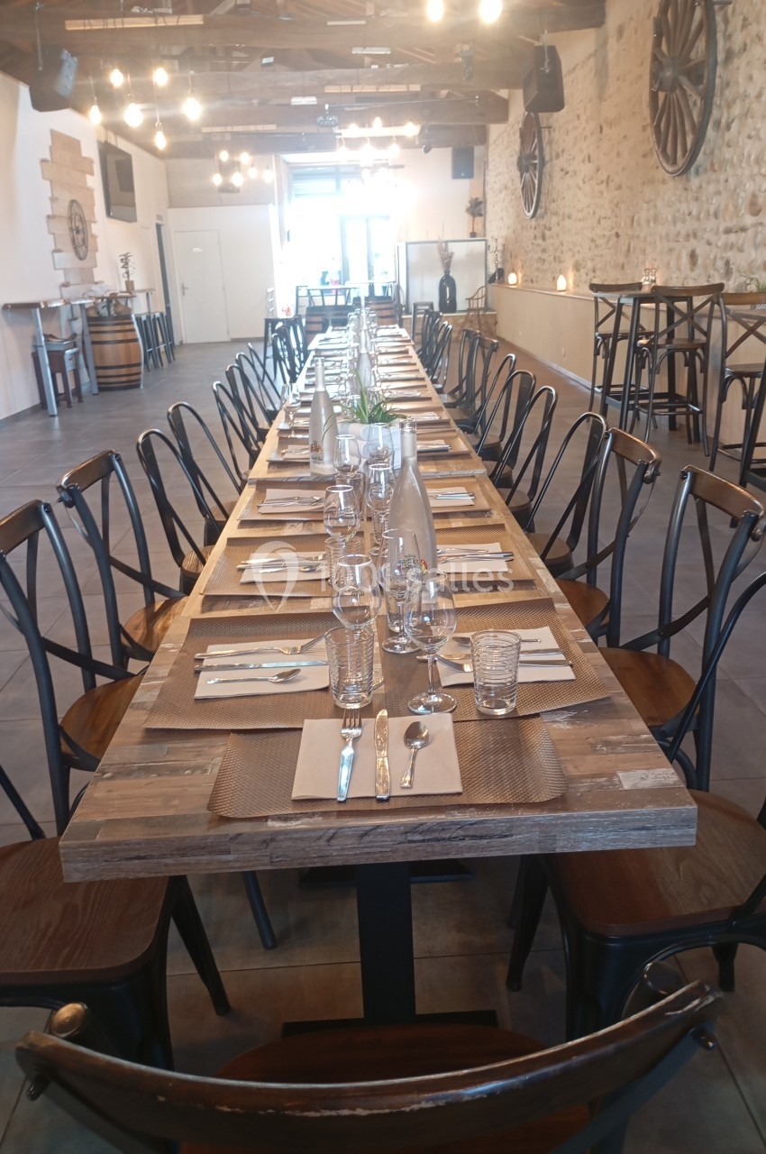 Grande table en bois dressée pour un repas, entourée de chaises, dans une salle au décor rustique avec murs en pierre.