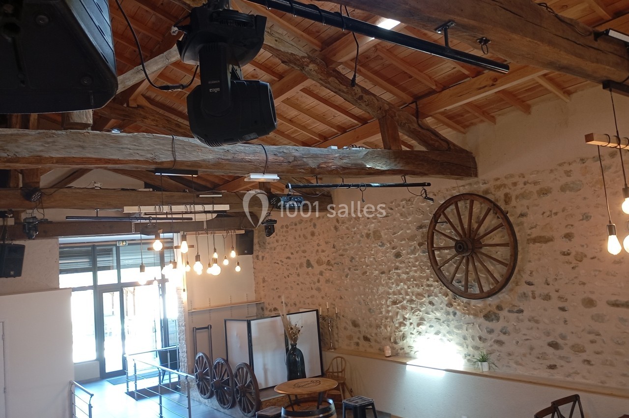 Salle avec murs en pierre, poutres apparentes, éclairage suspendu et décoration rustique, dont une roue en bois.