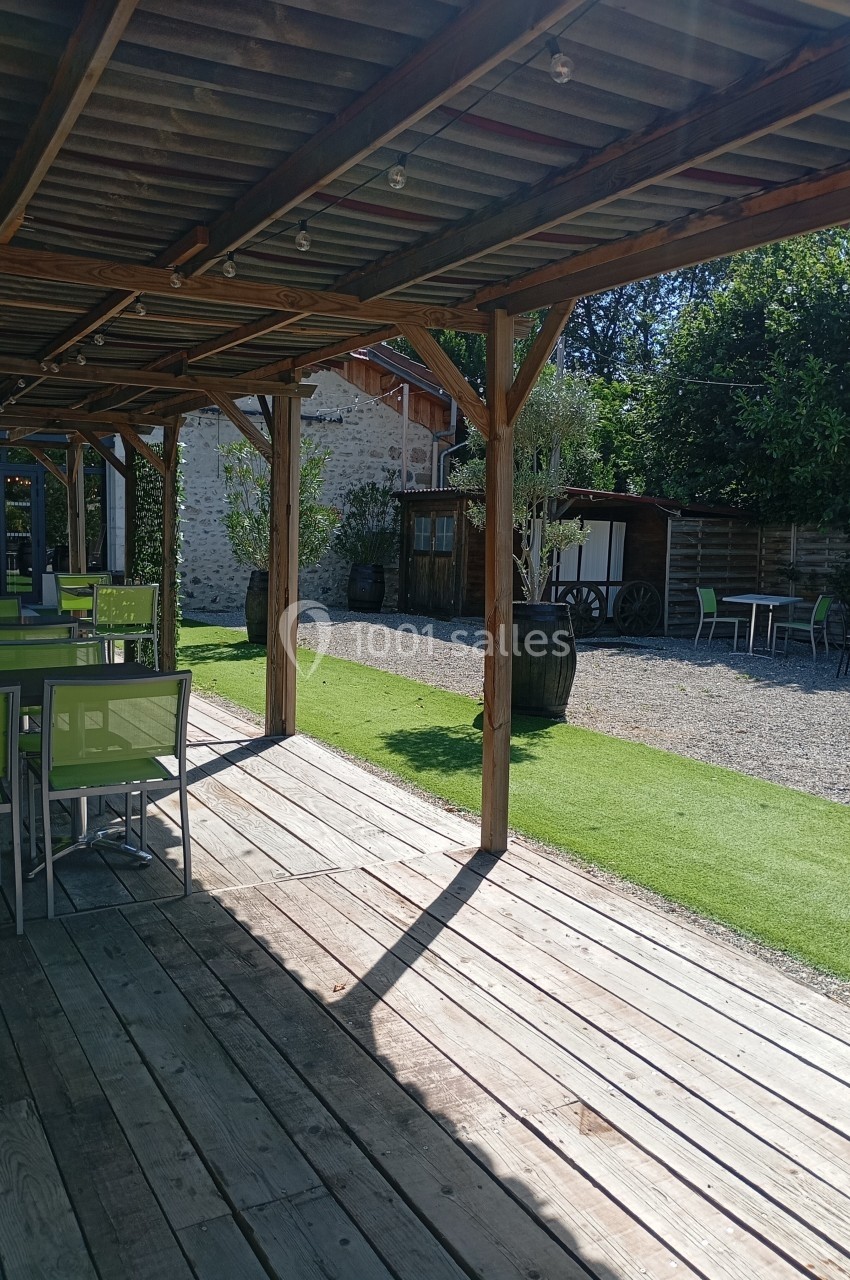 Terrasse en bois avec tables et chaises vertes, ombragée par une pergola, donnant sur une cour avec végétation.