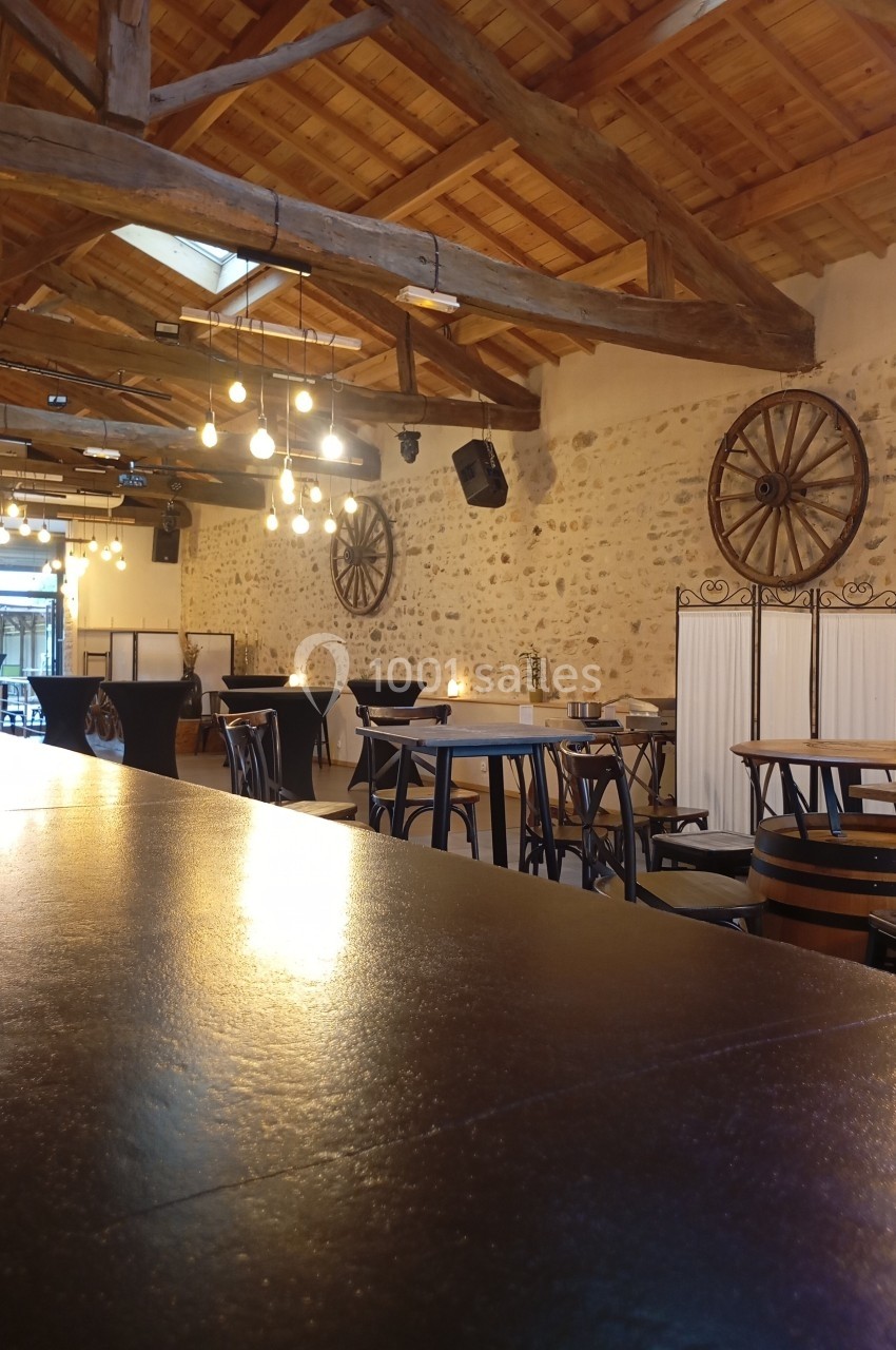Salle avec murs en pierre, poutres apparentes, tables en bois et éclairage suspendu, ambiance chaleureuse.