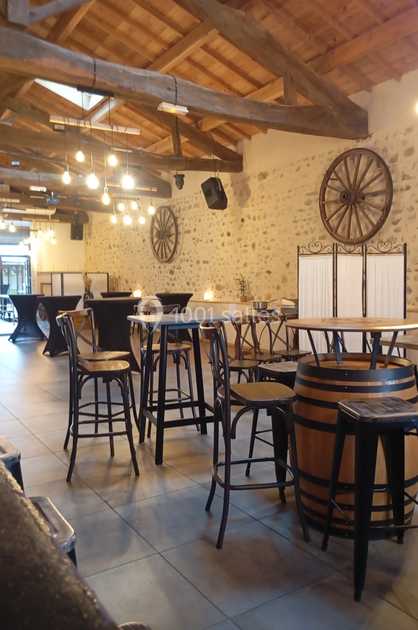 Salle avec murs en pierre, poutres apparentes, tables hautes et décoration rustique, éclairée par des suspensions.