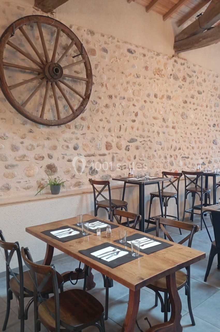 Salle de restaurant avec tables en bois, chaises en métal et mur en pierre décoré d'une roue en bois.