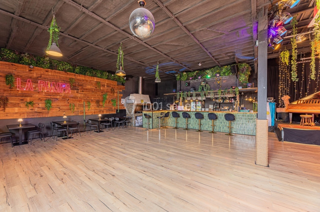 Salle spacieuse avec bar en bambou, tables, décorations végétales et enseigne lumineuse ’La Pampa’.