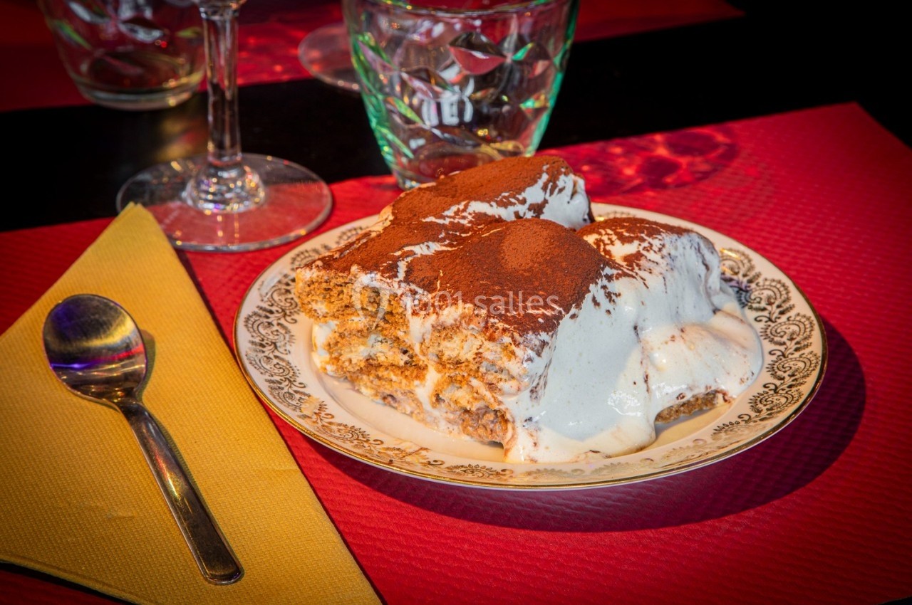 Assiette de tiramisu saupoudré de cacao, servie sur une table avec une cuillère et un verre en arrière-plan.