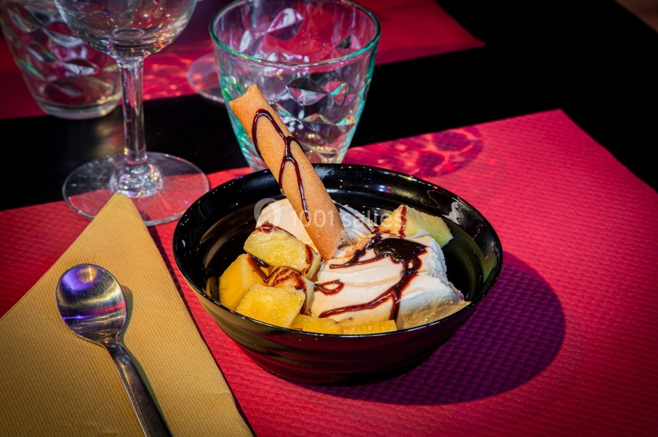 Coupe de dessert avec morceaux de fruits, glace, sauce chocolat et biscuit, posée sur une table avec serviette et verre.
