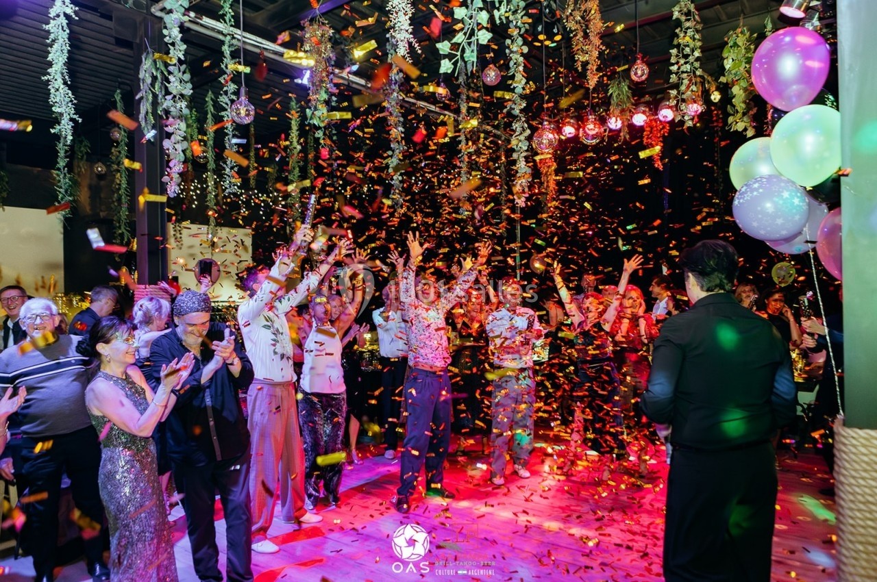 Groupe de personnes célébrant sous des confettis colorés dans une salle décorée avec des plantes suspendues et des ballons.