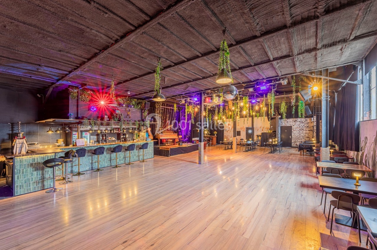Salle spacieuse avec bar, tables, éclairage coloré et décorations végétales suspendues.