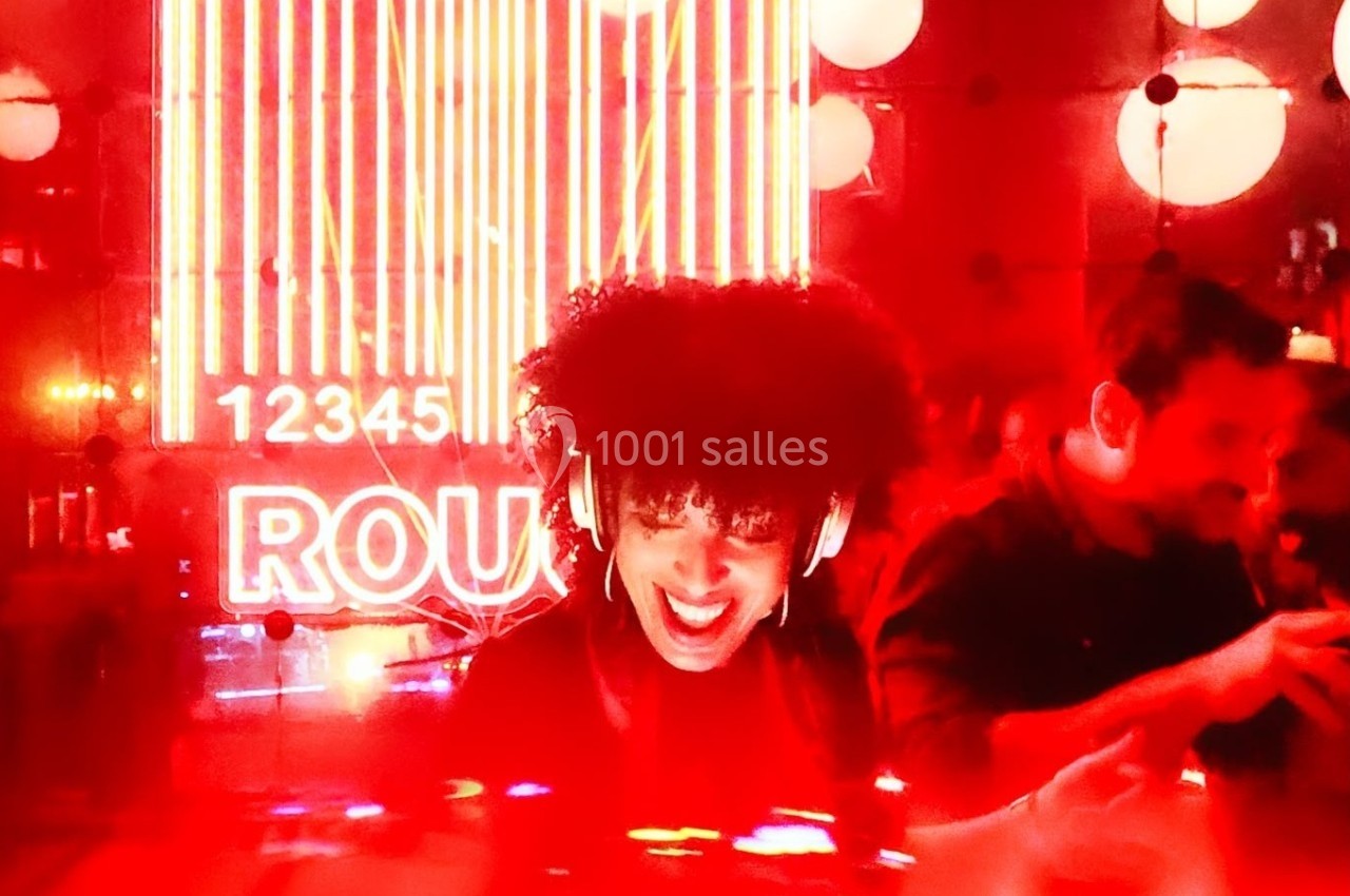 Une DJ souriante mixe dans une ambiance lumineuse rouge avec des néons et un public en arrière-plan.