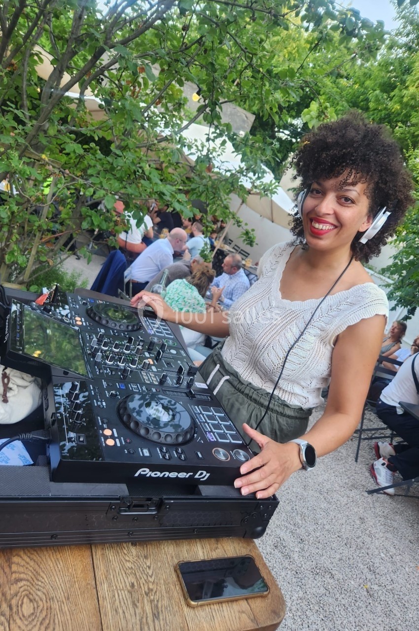 Une DJ souriante mixe de la musique en plein air lors d'un événement, entourée de verdure et de participants.