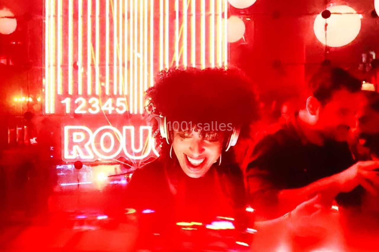 Une DJ souriante mixe dans une ambiance lumineuse rouge, avec un néon et des chiffres en arrière-plan.