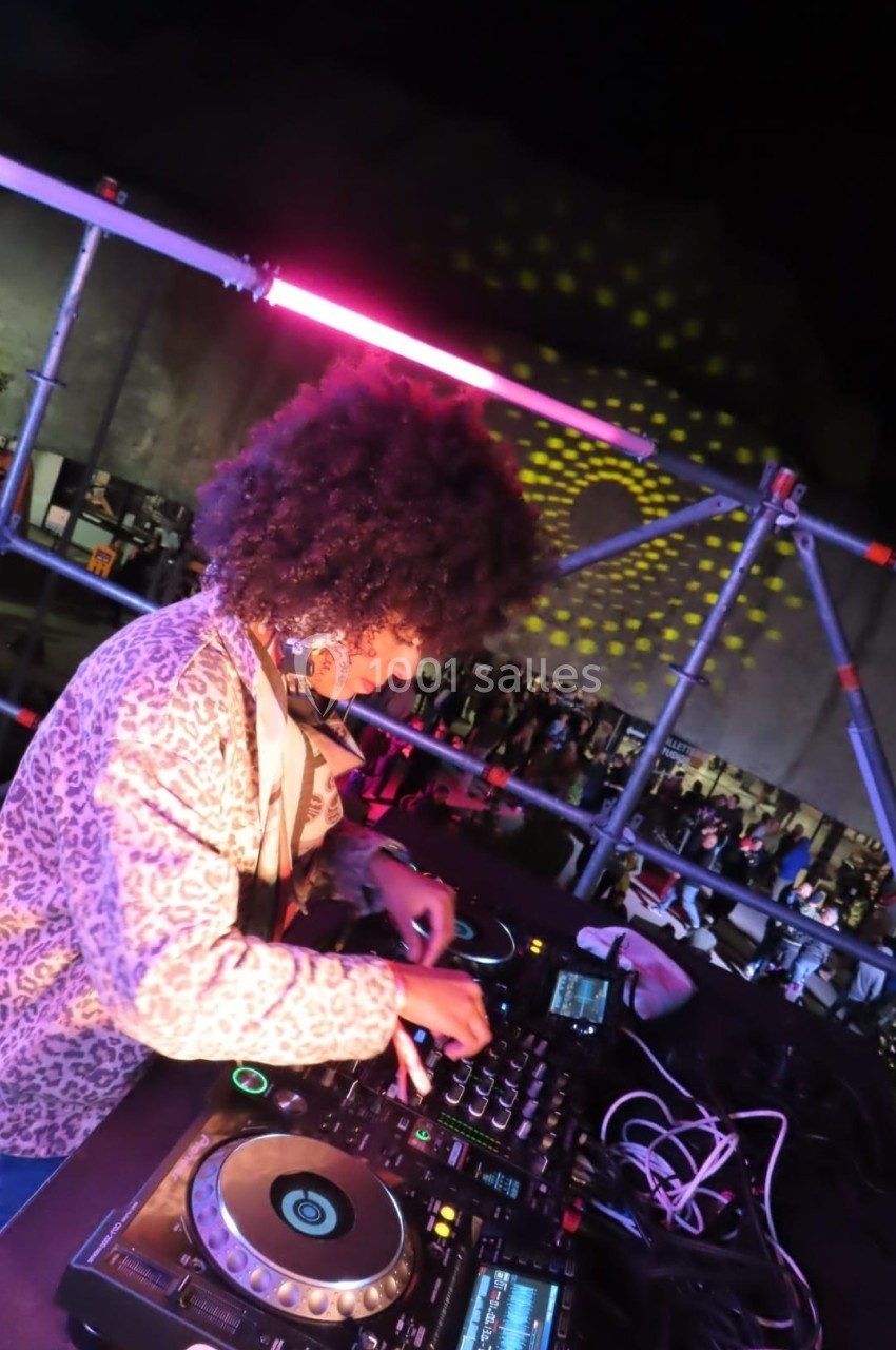 DJ mixant sur une scène en plein air, éclairée par des lumières colorées, avec un public en arrière-plan.