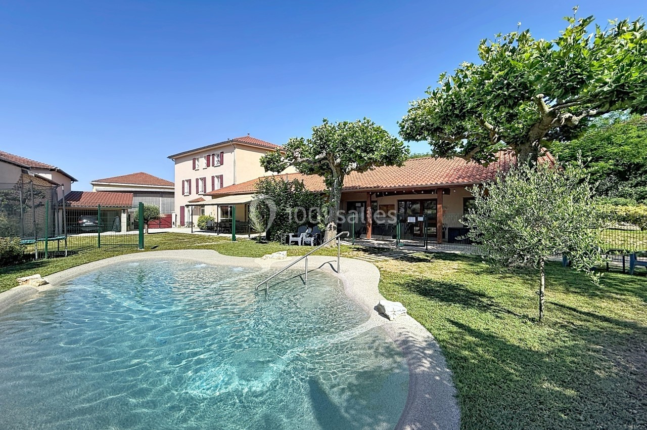 Piscine extérieure peu profonde entourée de verdure, avec une maison à deux étages en arrière-plan.