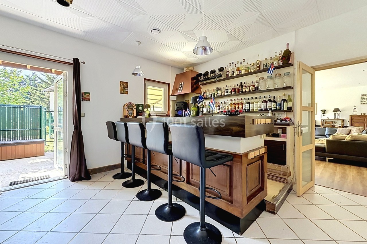 Bar intérieur avec comptoir en bois, tabourets noirs, étagères de bouteilles et porte ouverte sur un salon lumineux.