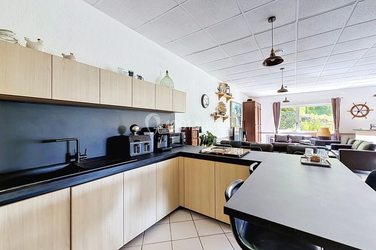 Cuisine moderne avec meubles en bois clair, plan de travail noir, et vue sur un salon lumineux avec grandes fenêtres.