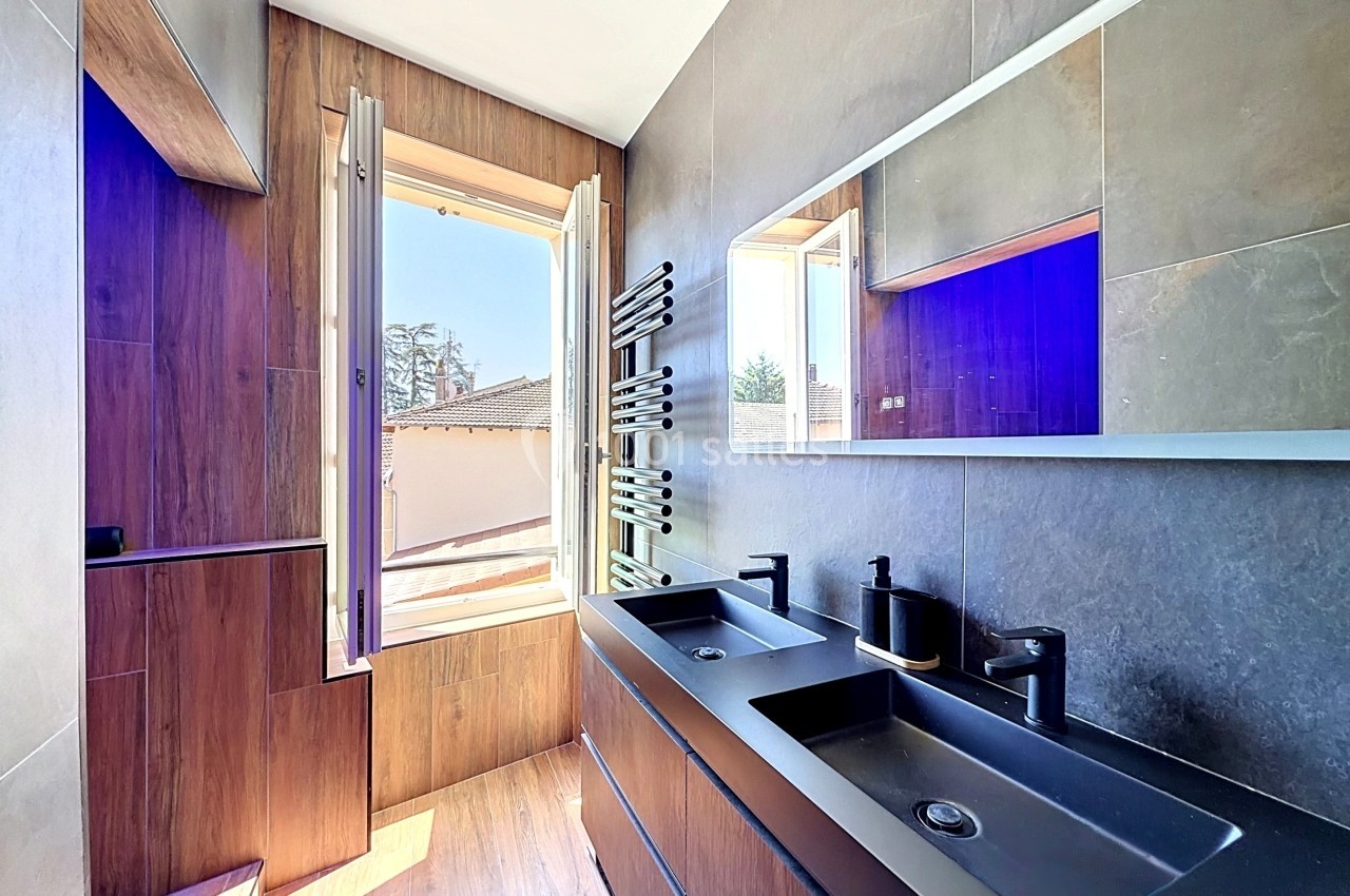 Salle de bain moderne avec deux vasques noires, miroir éclairé, fenêtre ouverte et murs en bois et carrelage.