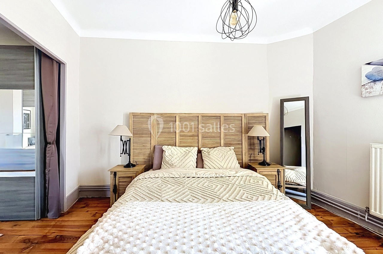 Chambre lumineuse avec lit double, tête de lit en bois, lampes de chevet et miroir sur pied, parquet au sol.
