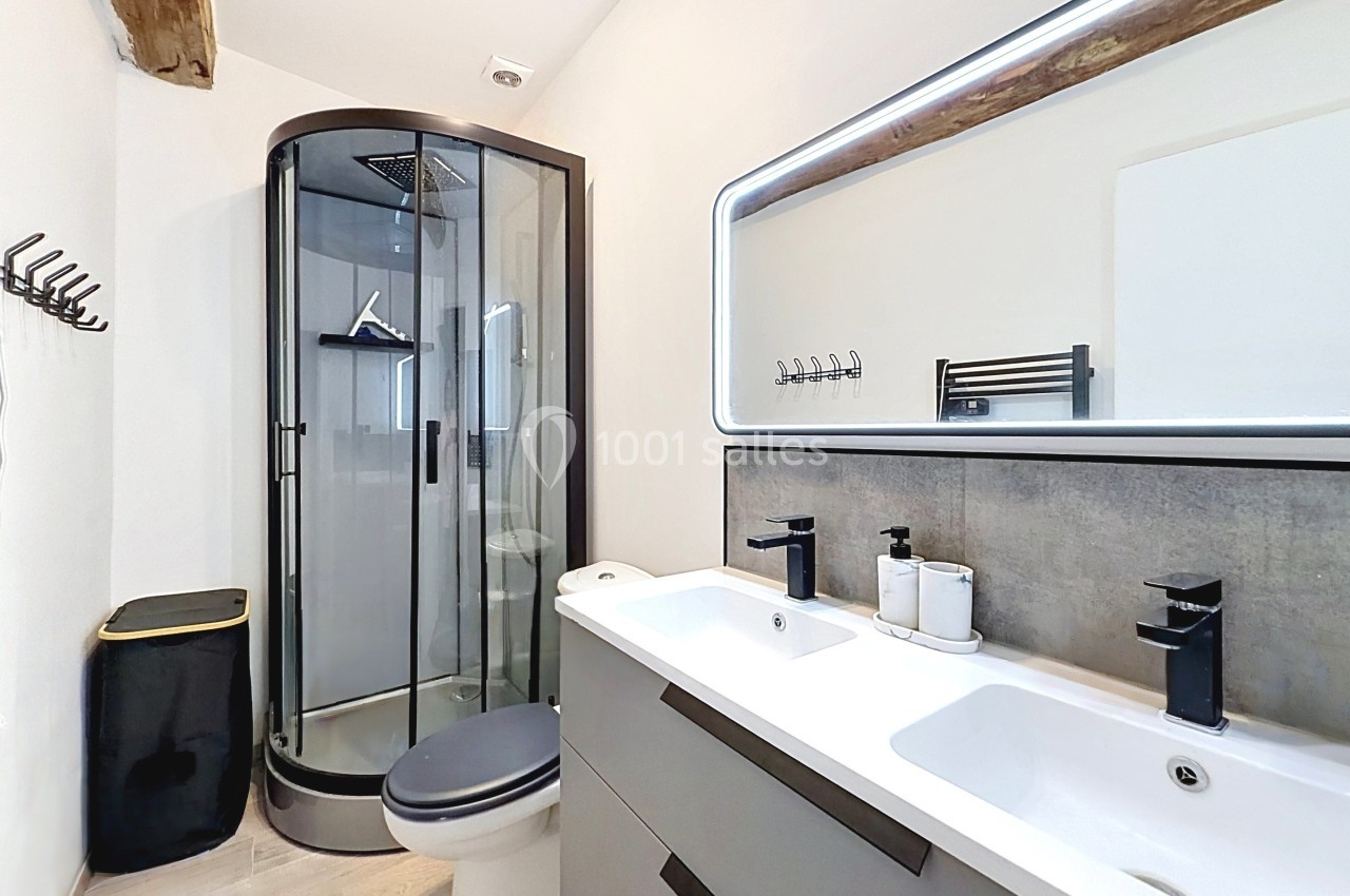 Salle de bain moderne avec douche arrondie, double vasque, miroir éclairé et poutre apparente au plafond.
