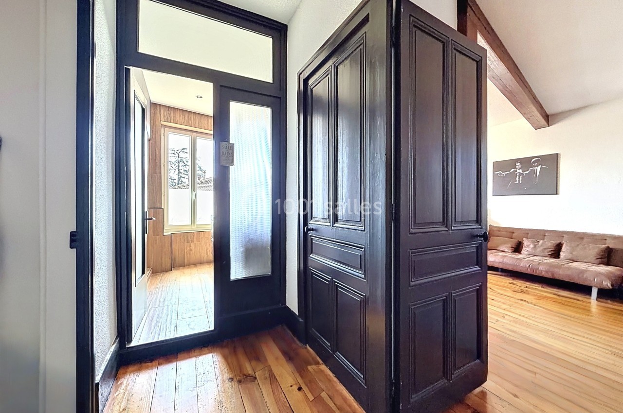 Vue d'un couloir lumineux avec parquet en bois, portes noires et accès à un salon et une pièce vitrée.