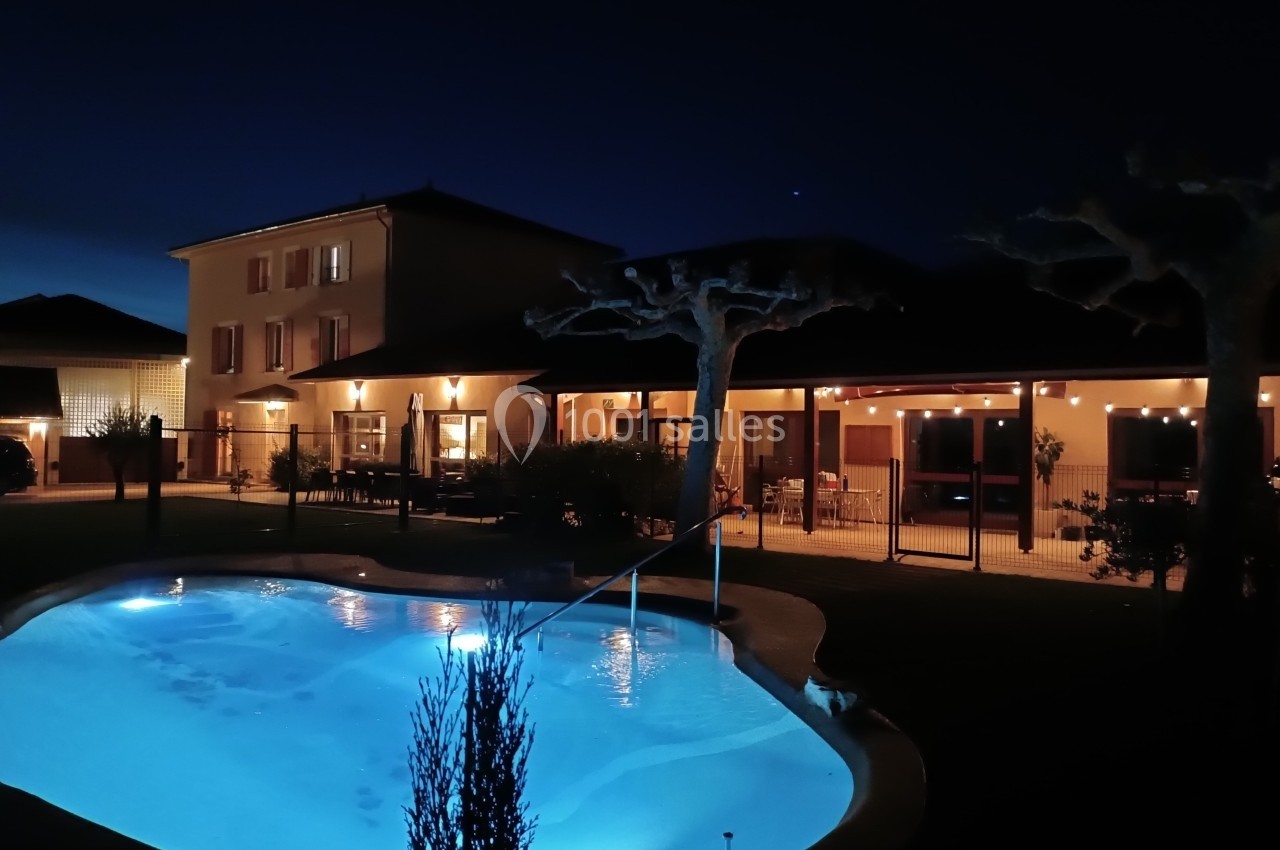 Maison éclairée de nuit avec une piscine illuminée au premier plan et une terrasse aménagée en arrière-plan.