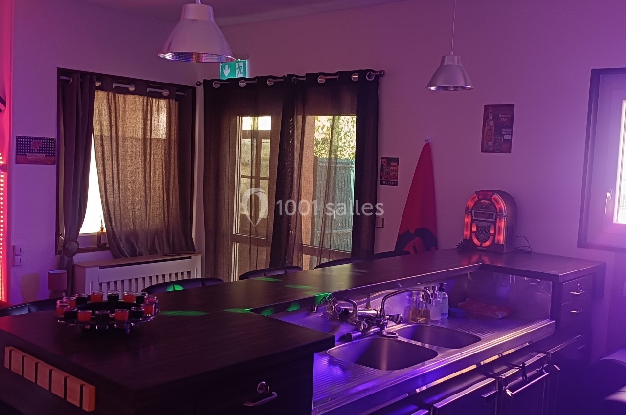 Cuisine moderne avec un bar central en bois, éclairage coloré et rideaux fermés devant des portes vitrées.