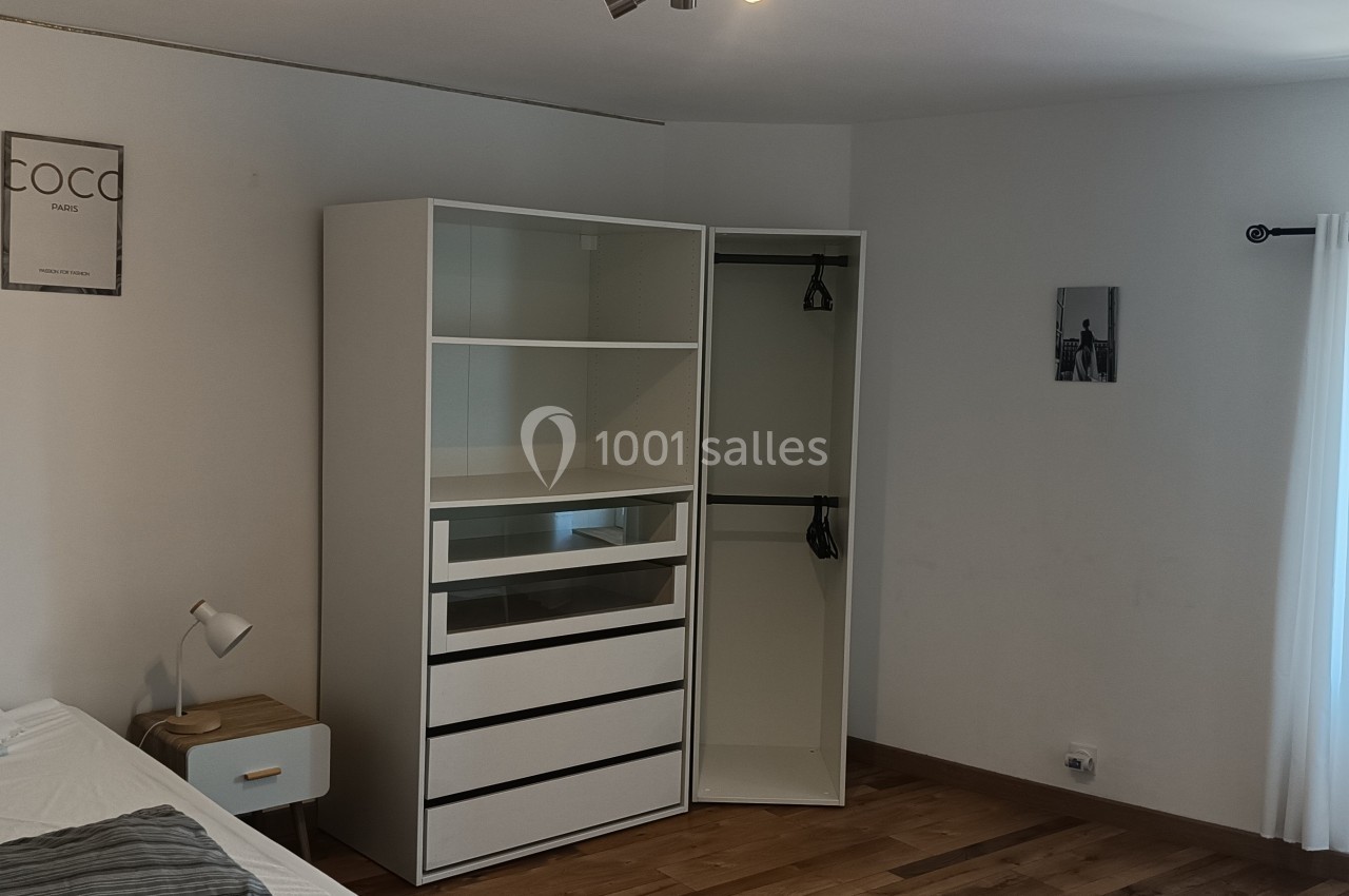 Chambre lumineuse avec lit, table de chevet, armoire ouverte blanche et fenêtre donnant sur un paysage extérieur.