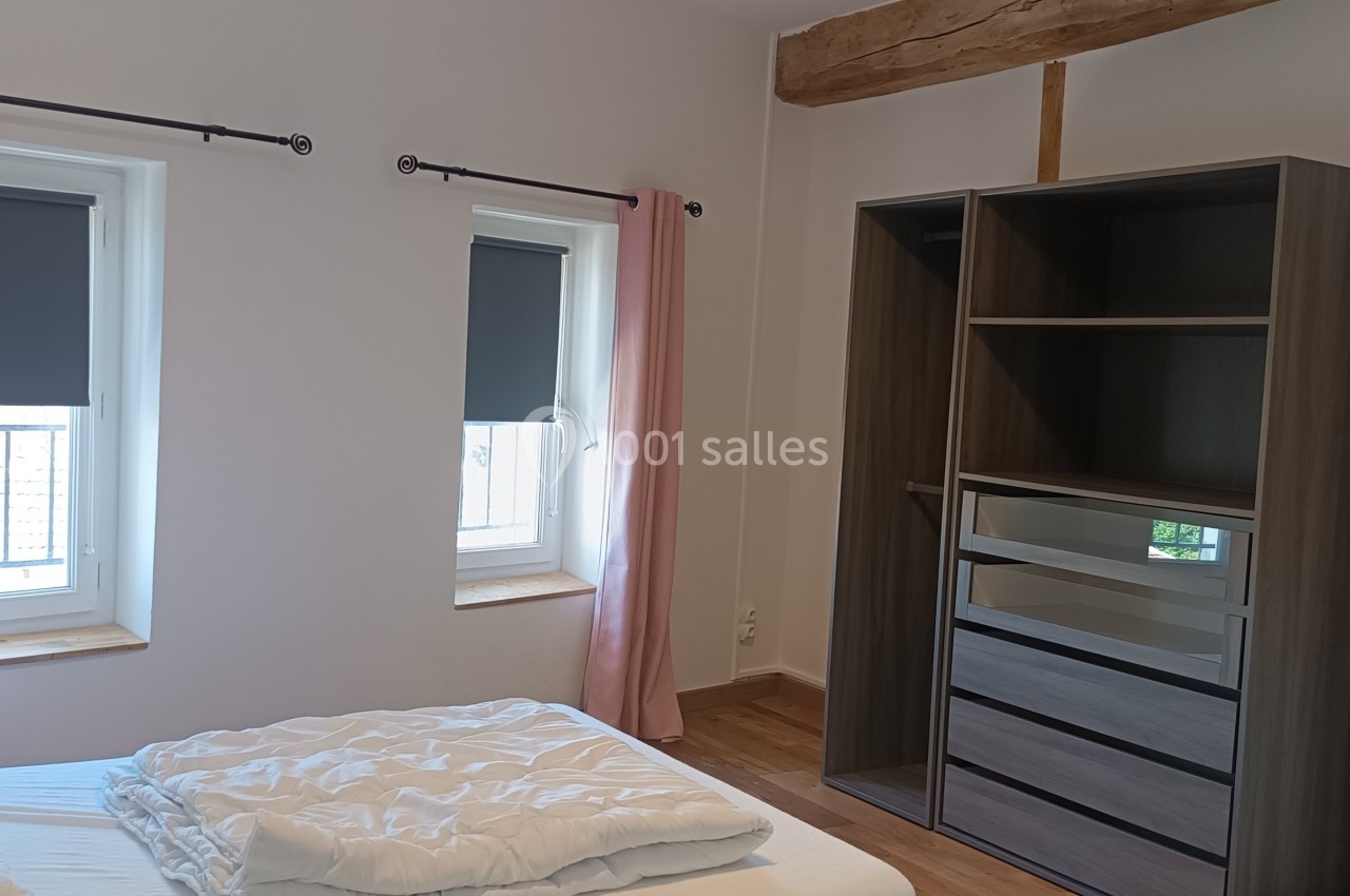Chambre lumineuse avec lit double, armoire en bois sombre, deux fenêtres avec rideaux roses et volets roulants.