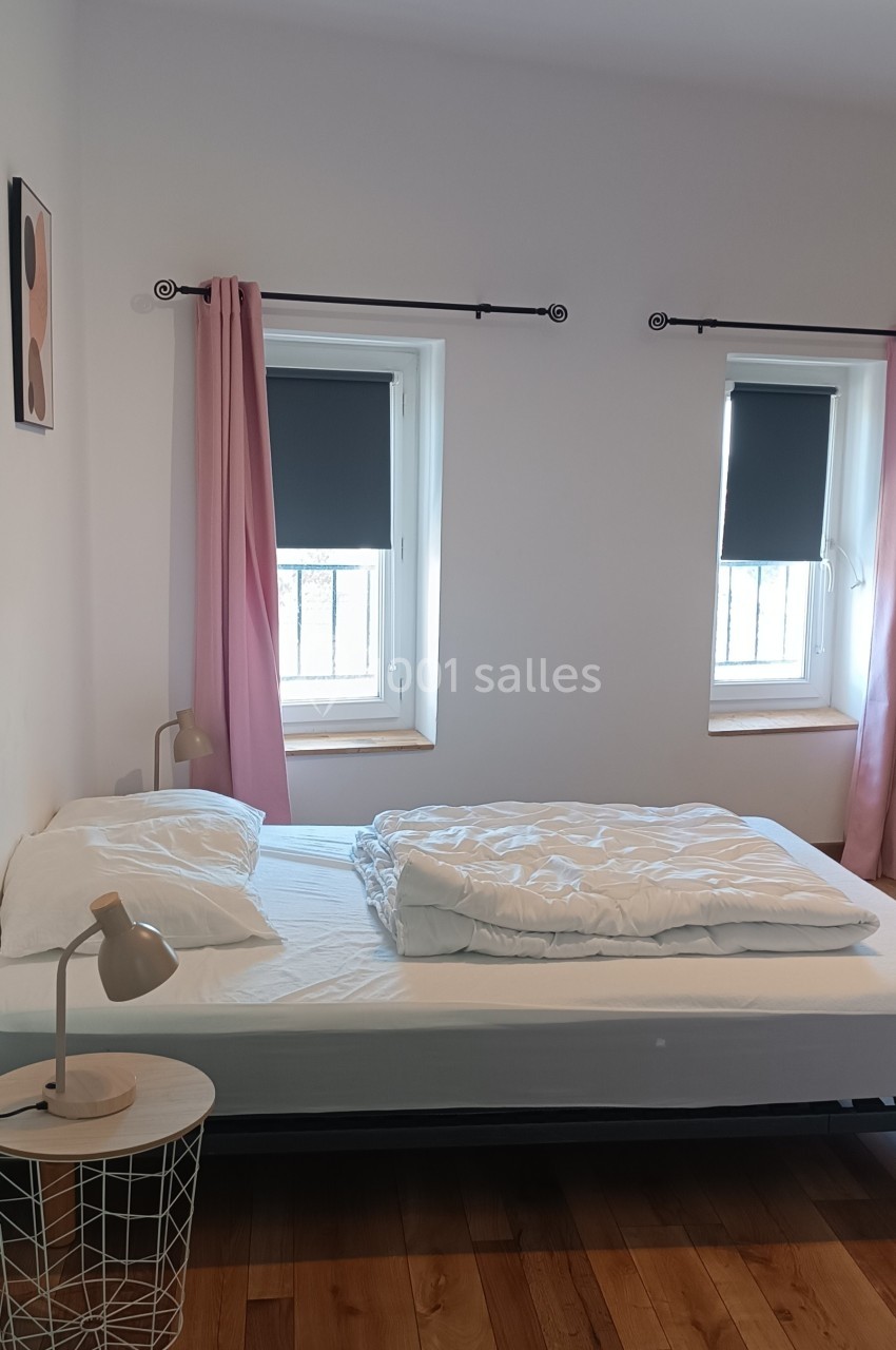 Lit simple avec draps blancs, table de chevet avec lampe, parquet en bois et deux fenêtres avec rideaux roses.