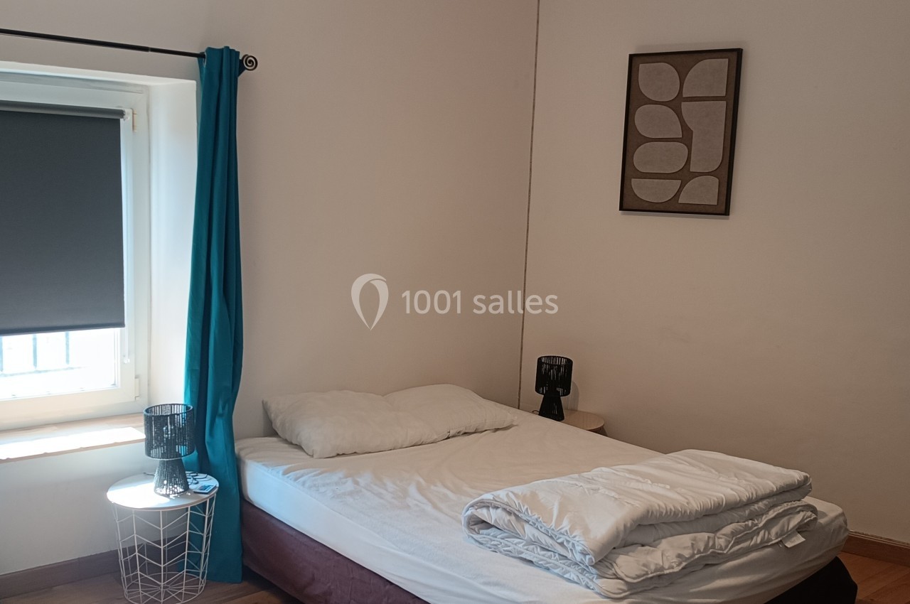 Chambre simple avec un lit, une fenêtre avec rideaux bleus et stores, deux lampes de chevet et des cadres au mur.