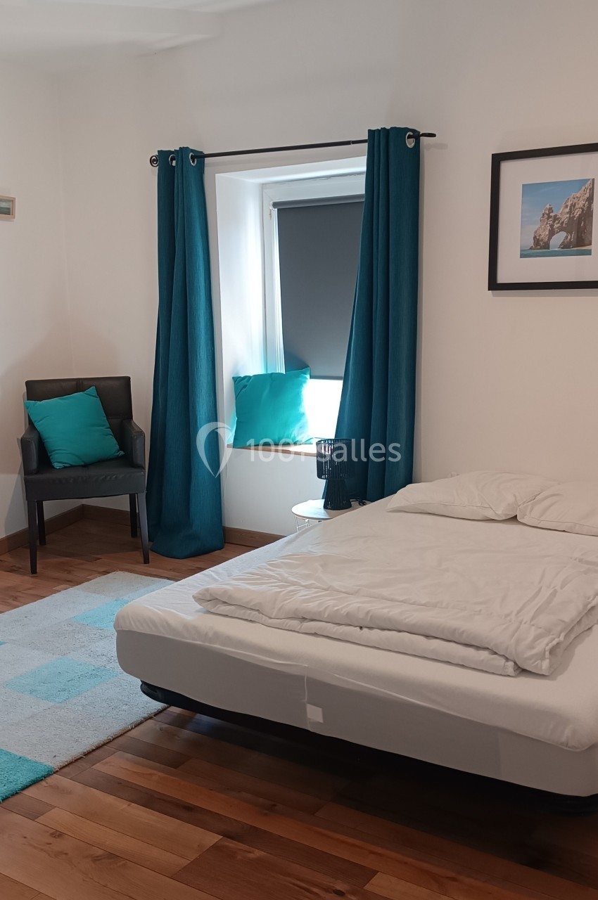 Chambre lumineuse avec lit double, parquet, fauteuil, rideaux bleus et fenêtre donnant sur l'extérieur.