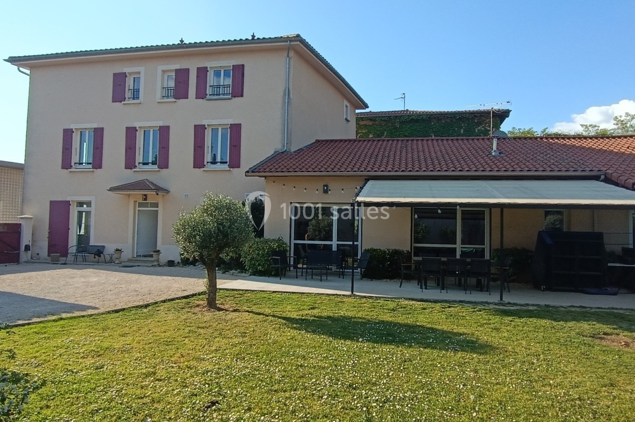 Maison avec façade beige, volets rouges, terrasse couverte et jardin arboré sous un ciel dégagé.