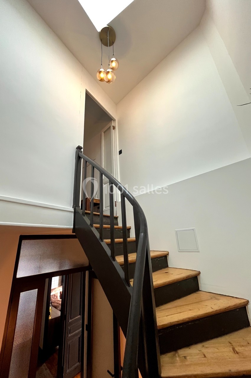 Escalier en bois avec rampe noire menant à un étage supérieur, éclairé par une suspension sous une lucarne.