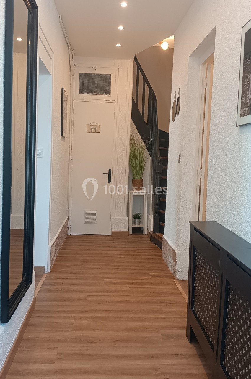 Couloir lumineux avec parquet, miroir mural, meuble noir, escalier en bois et plante verte au fond.