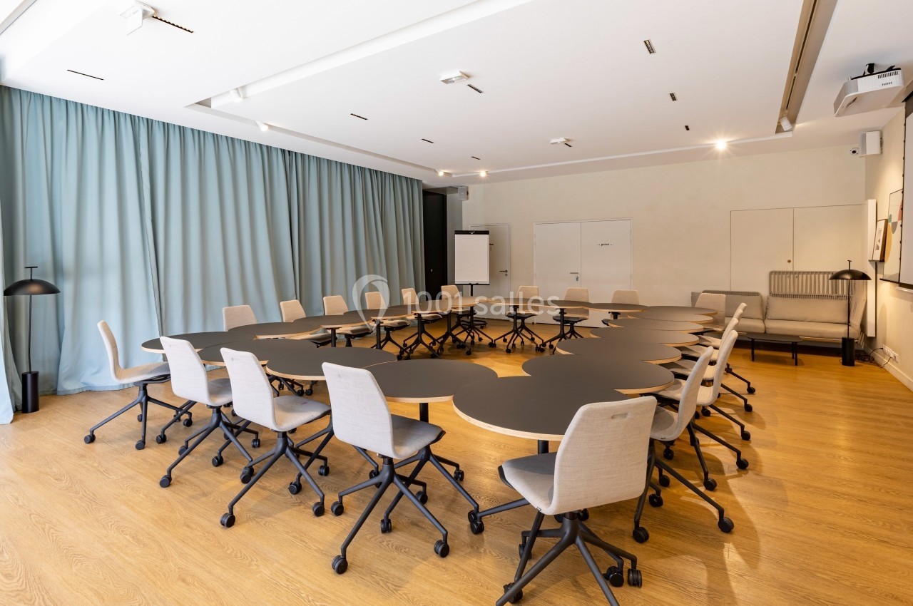 Salle de réunion lumineuse avec tables noires modulables, chaises à roulettes et rideaux bleus en arrière-plan.