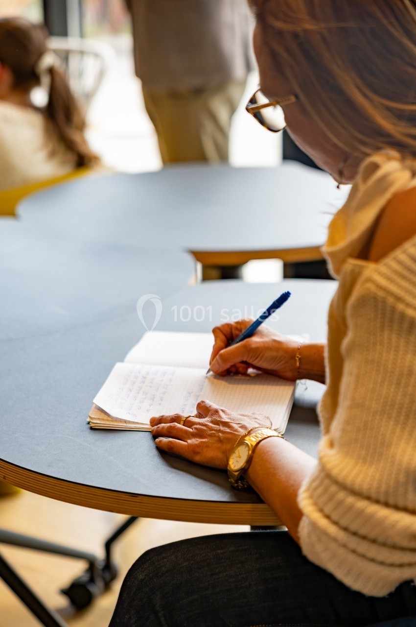 Femme écrivant dans un carnet à une table ronde, dans un environnement intérieur lumineux.