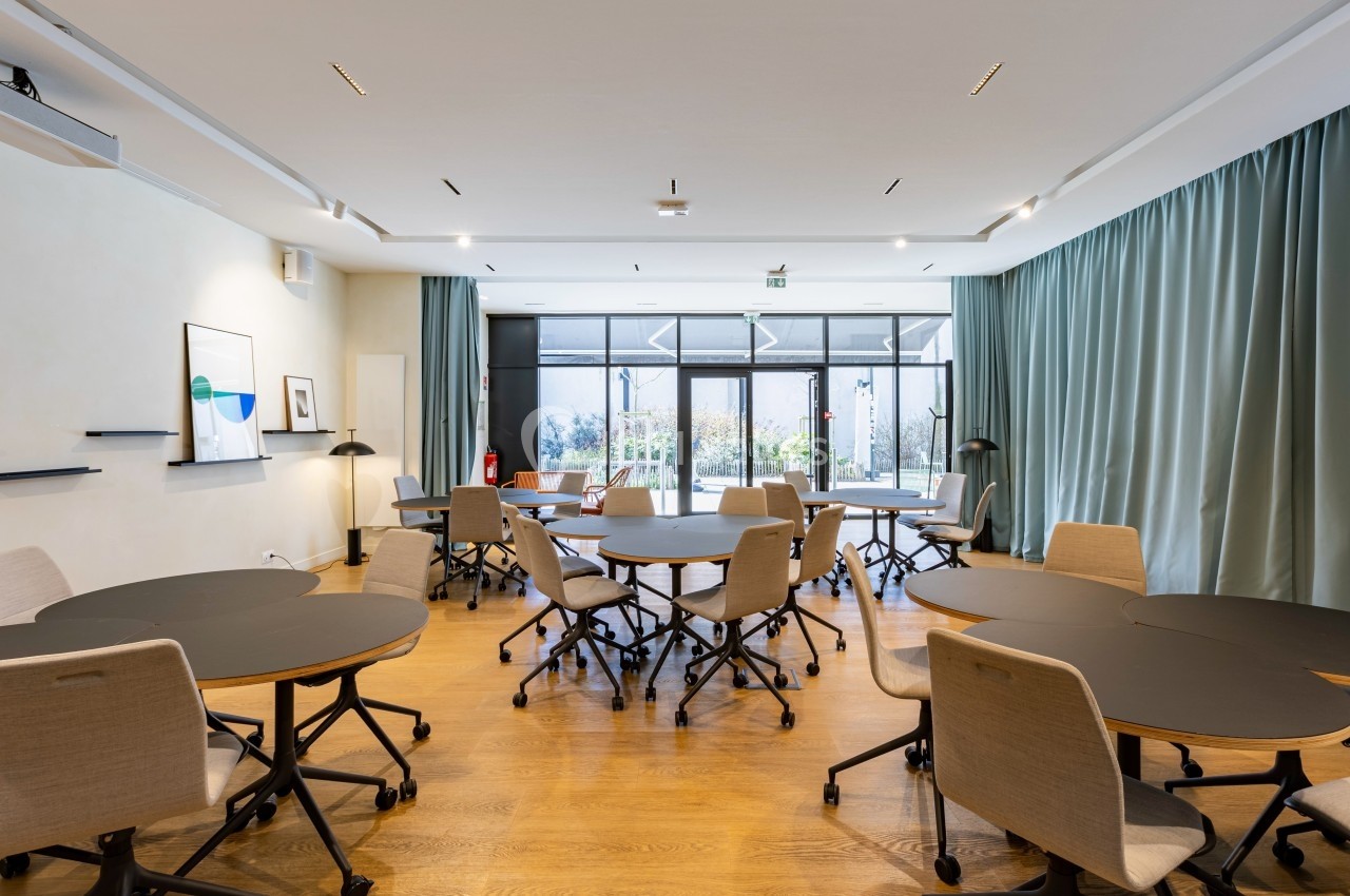 Salle lumineuse avec plusieurs tables rondes noires entourées de chaises, grandes baies vitrées et rideaux verts.