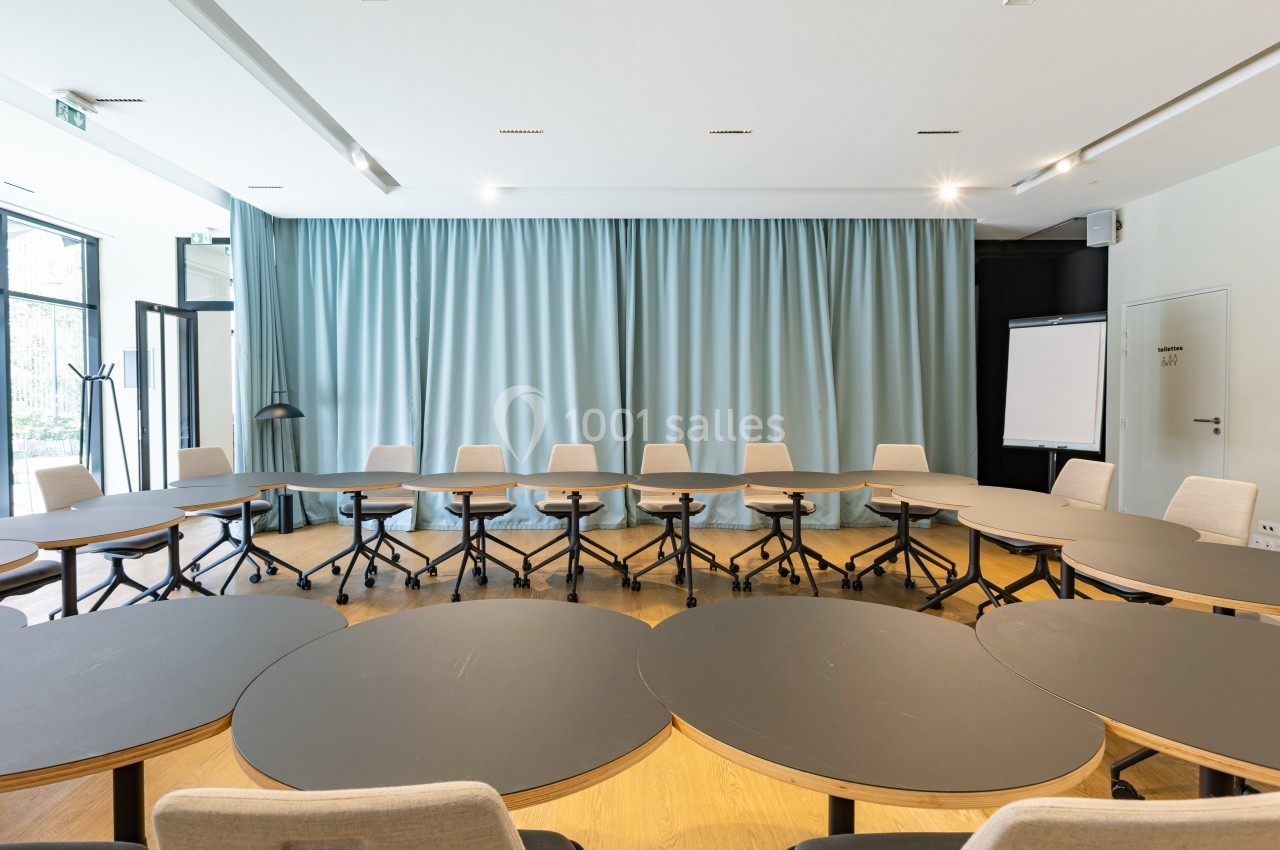 Salle de réunion lumineuse avec tables ovales noires, chaises à roulettes et rideaux bleus en arrière-plan.