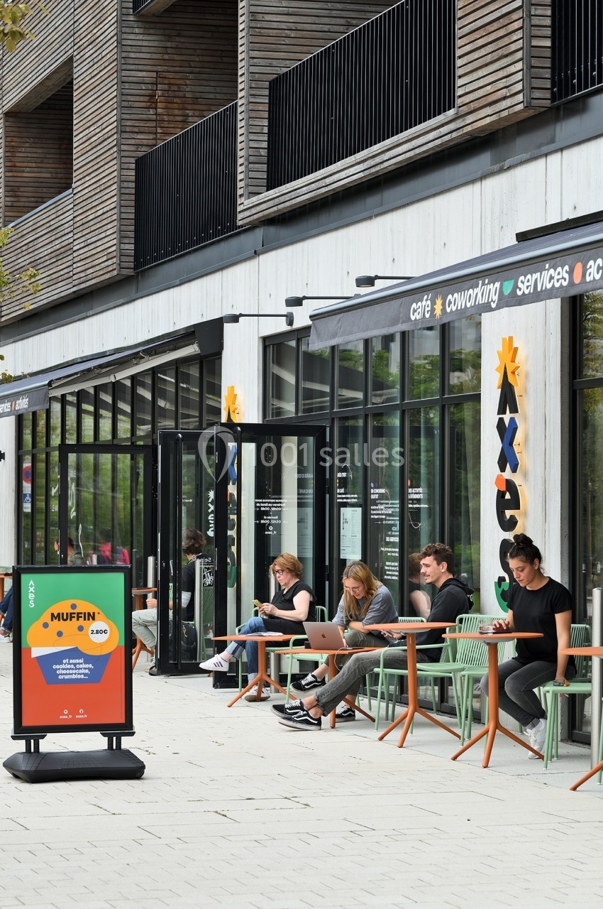 Personnes assises à des tables en terrasse d'un café moderne, travaillant ou discutant, avec une enseigne visible.