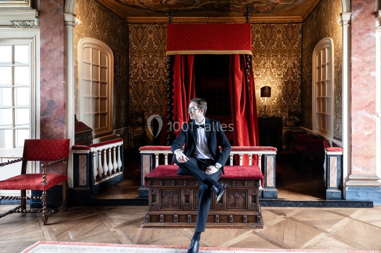Un homme en costume assis sur un banc en bois dans une pièce ornée de décorations historiques et d'un lit à baldaquin rouge.