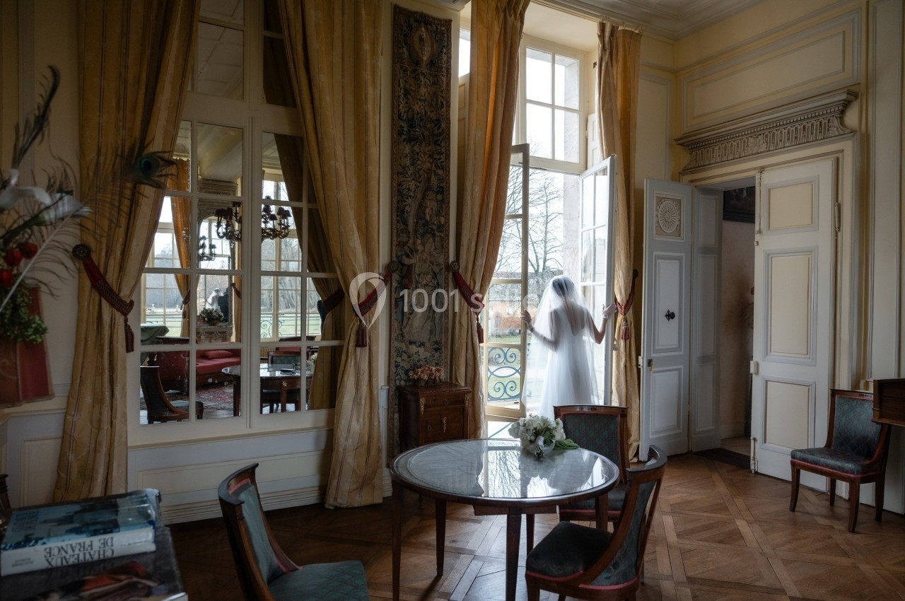 Une personne en tenue claire se tient près d'une porte-fenêtre ouverte dans une pièce élégante au décor classique.