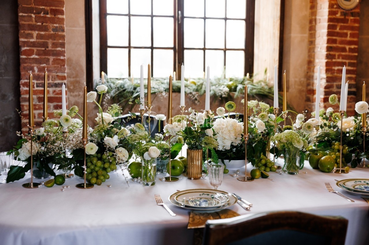 Table décorée avec des fleurs blanches, des bougies dorées et des fruits verts, devant une fenêtre à carreaux.