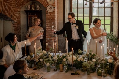 Des mariés et leurs invités trinquent autour d'une table décorée de fleurs et de bougies dans une salle rustique.