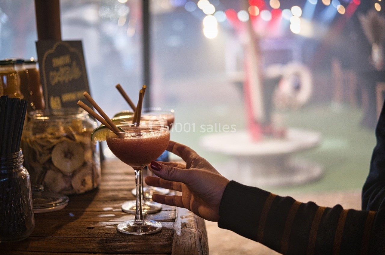 Main tenant un verre de cocktail sur un comptoir en bois, avec un décor festif et lumineux en arrière-plan.