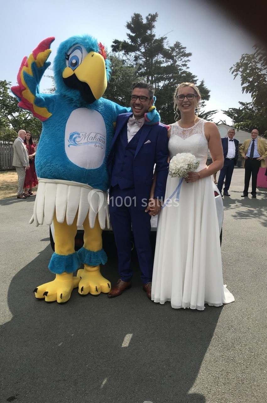 Un couple de mariés souriant pose à l'extérieur avec une mascotte en forme d'oiseau bleu.